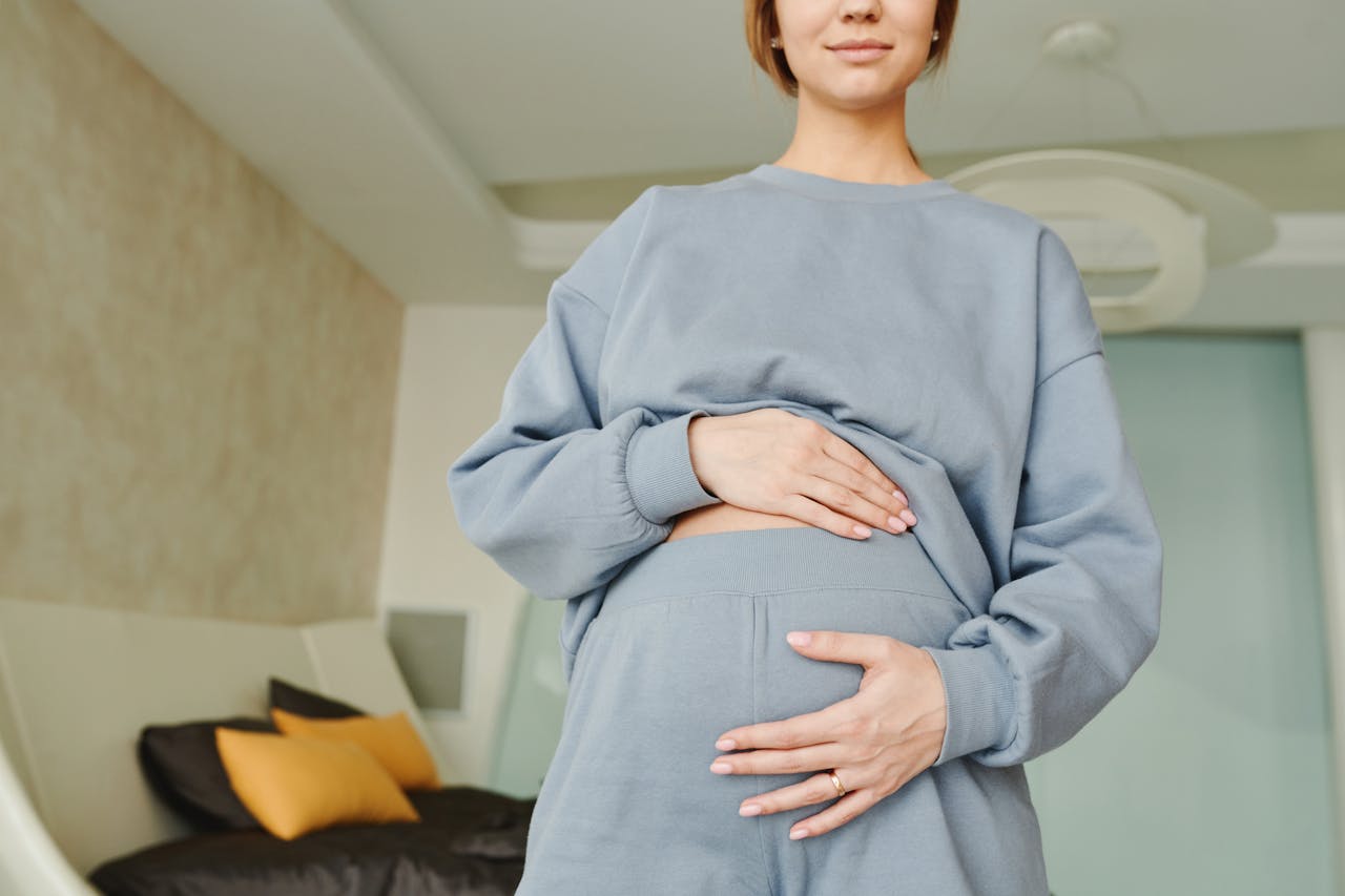 A close-up of a pregnant woman indoors, wearing a blue sweater, hand resting on her belly.